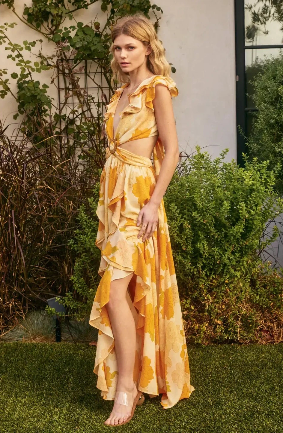 Woman wearing a yellow floral dress standing outdoors with greenery around.