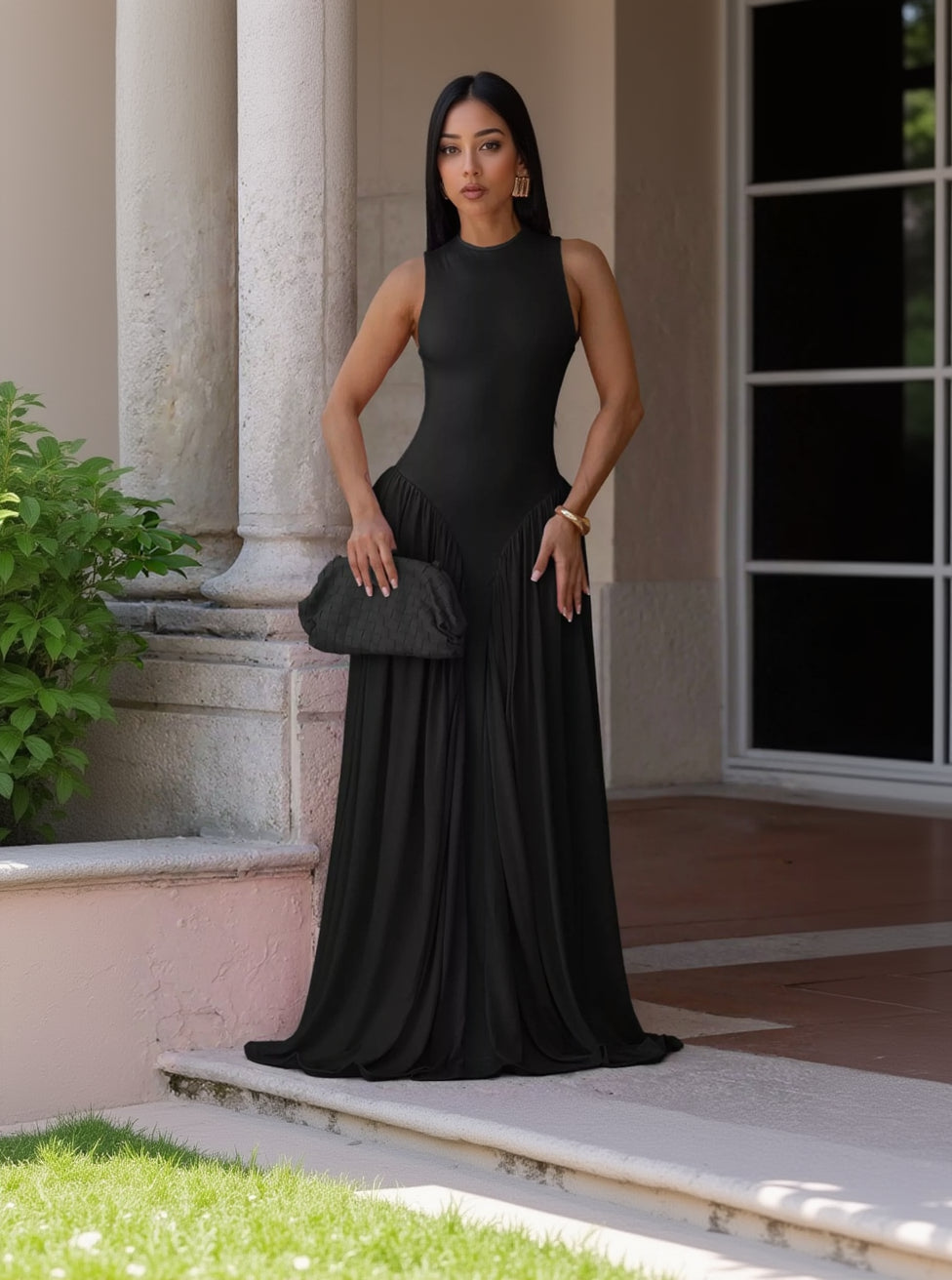 A woman wearing a sleeveless black maxi dress with a high mock neck and pleated skirt, holding a black clutch bag while standing on a porch.