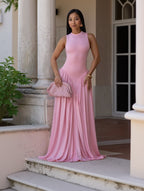 A woman wearing a sleeveless, high-neck, floor-length pink gown with a gathered, pleated skirt, holding a matching pink textured clutch bag, posed in front of a stone column.