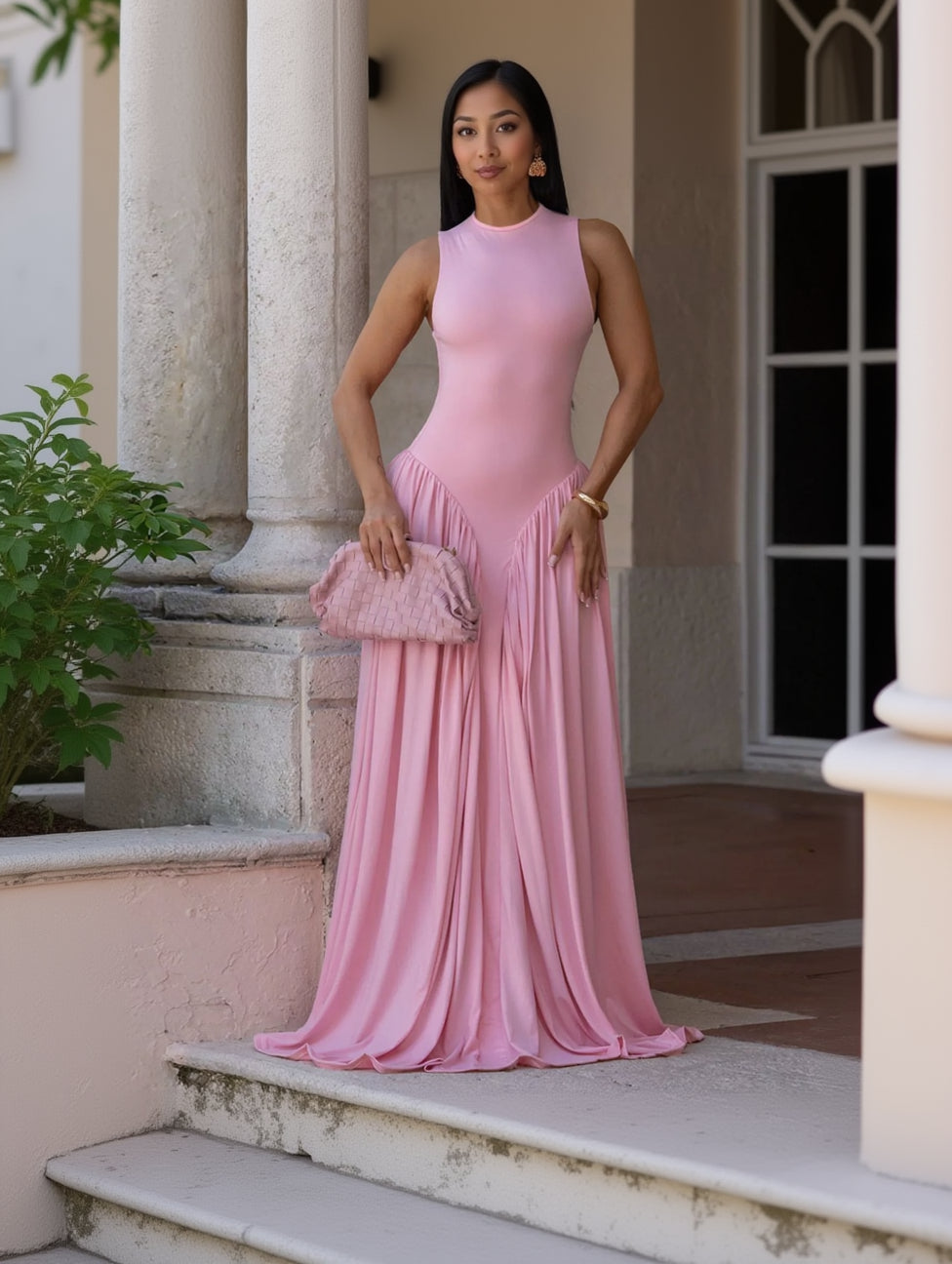 A woman wearing a sleeveless, high-neck, floor-length pink gown with a gathered, pleated skirt, holding a matching pink textured clutch bag, posed in front of a stone column.