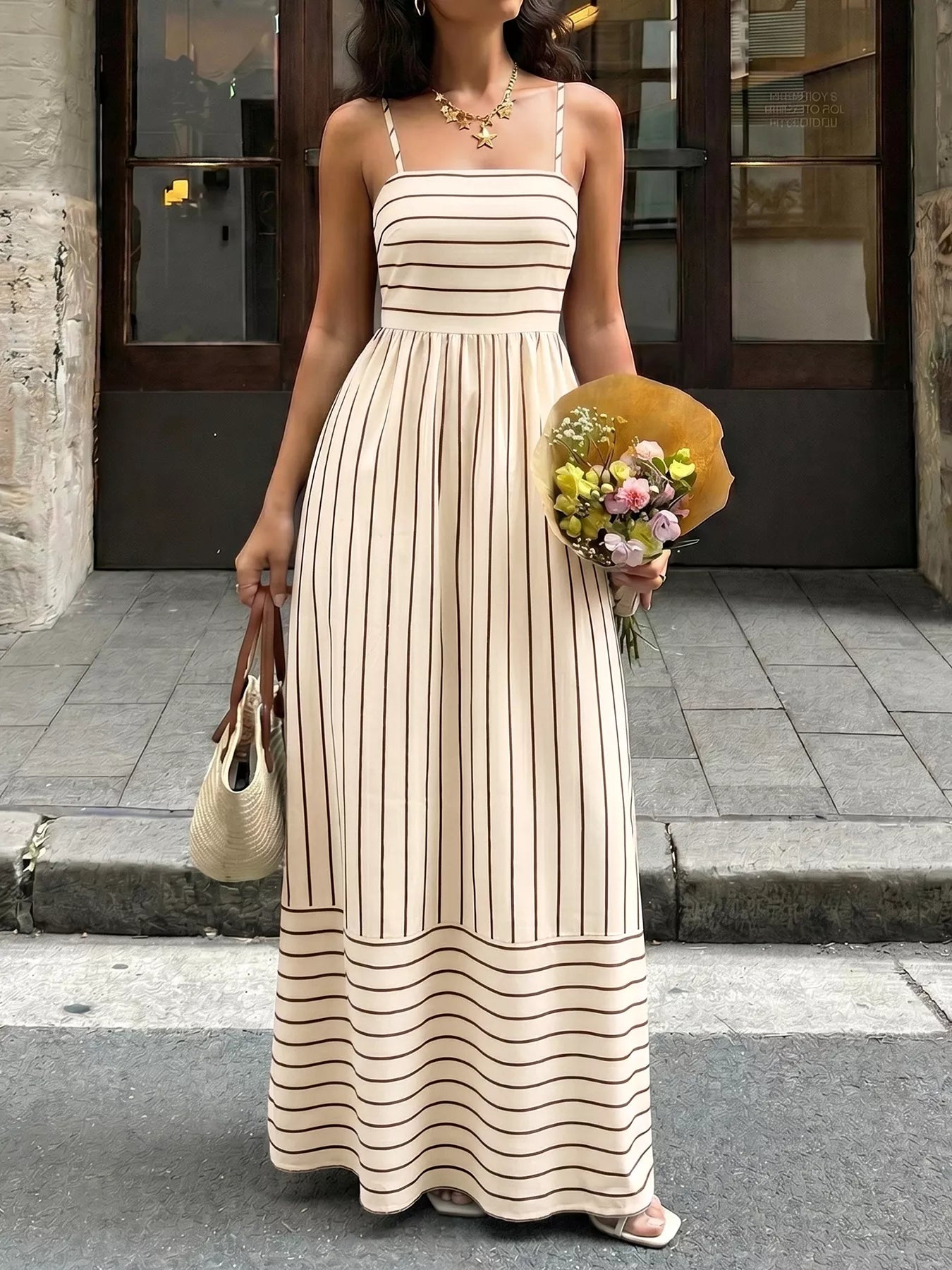 Woman in a long beige and black striped dress holding flowers and a bag on a city street.