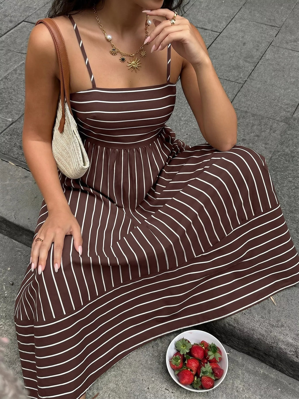Woman in a brown and white striped dress sitting on a stone surface with a bowl of strawberries.