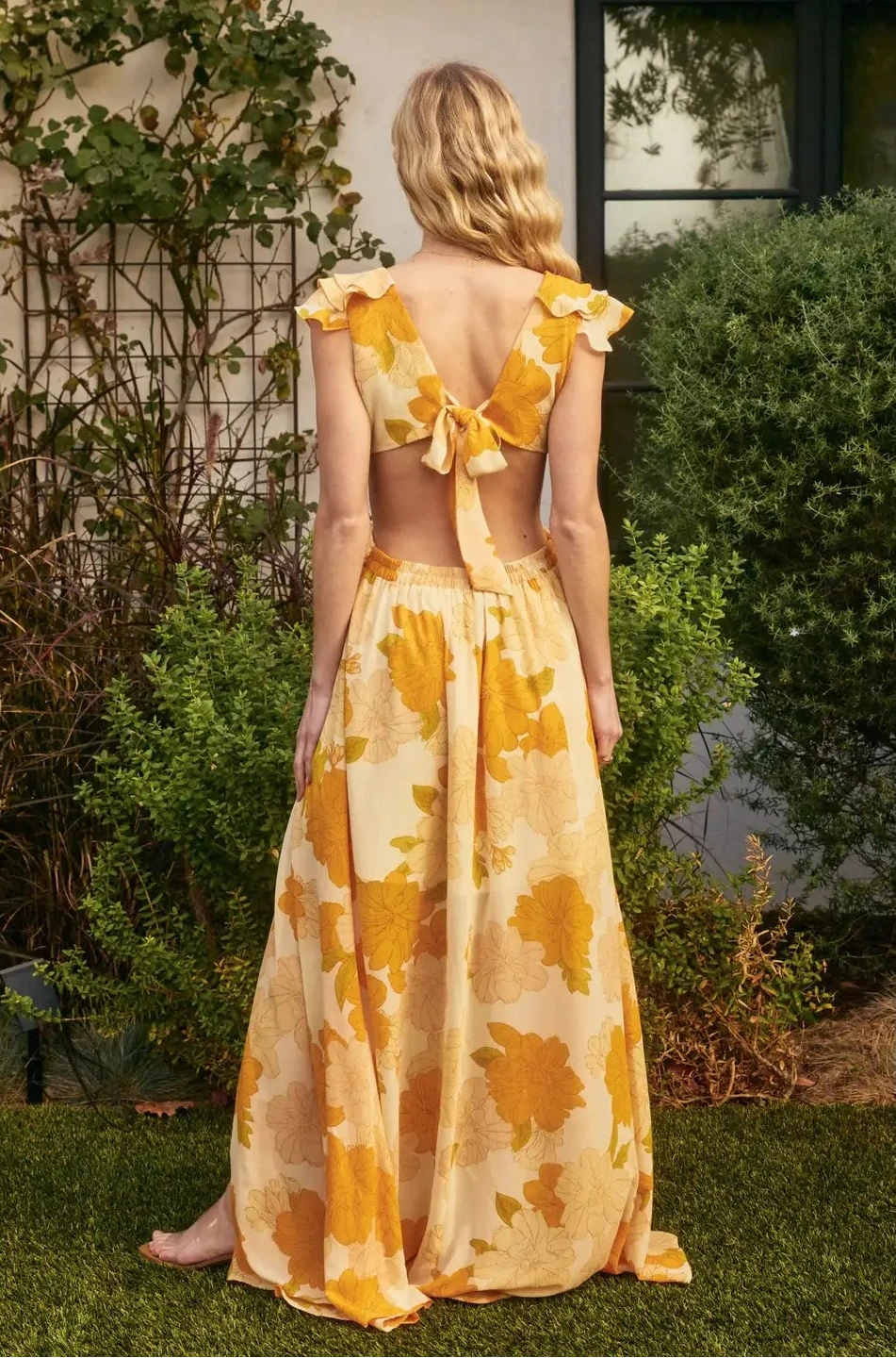 Woman wearing a yellow floral dress standing outdoors with greenery around.