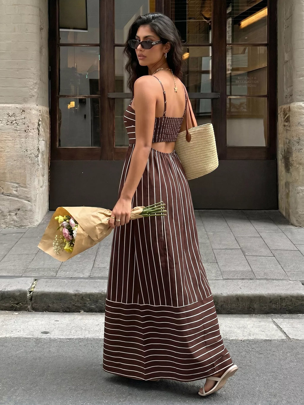 Woman in a long brown and white striped dress holding flowers and a straw bag on a city street.