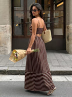 Woman in a long brown and white striped dress holding flowers and a straw bag on a city street.
