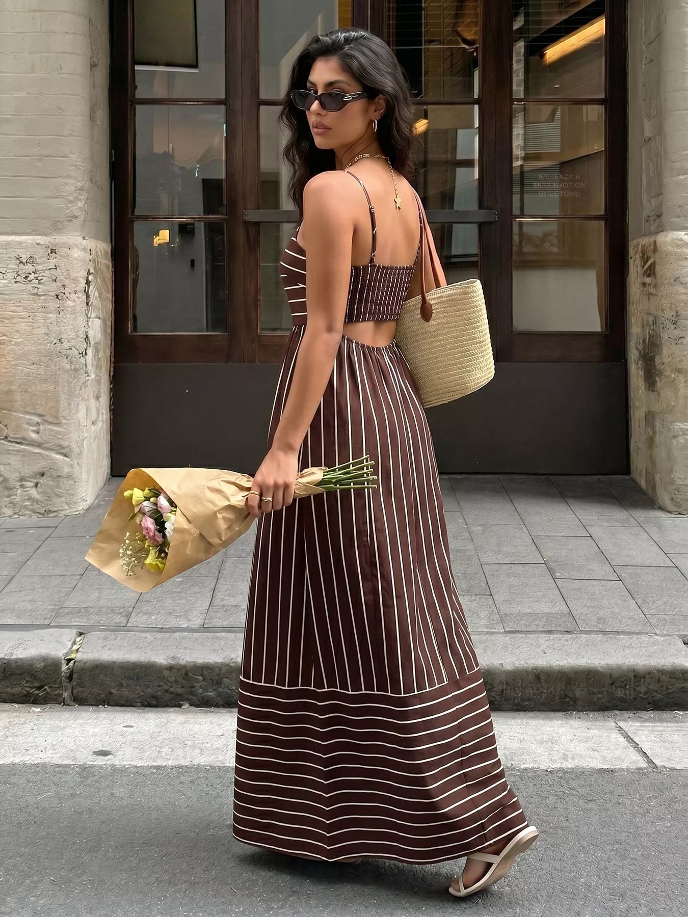 Woman in a long brown and white striped dress holding flowers and a straw bag on a city street.