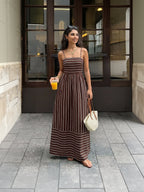 Woman in a brown and white striped dress holding a drink and a bag on a city street.