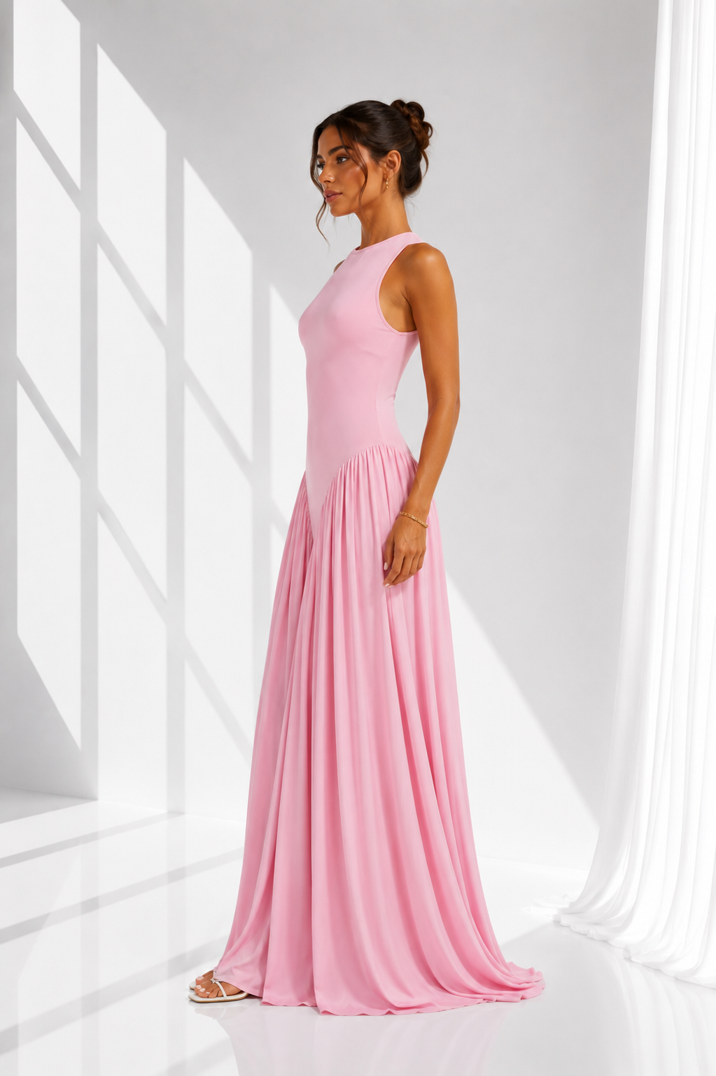 A woman modeling a sleeveless, floor-length pink gown in a bright white studio with dramatic window-like shadows.