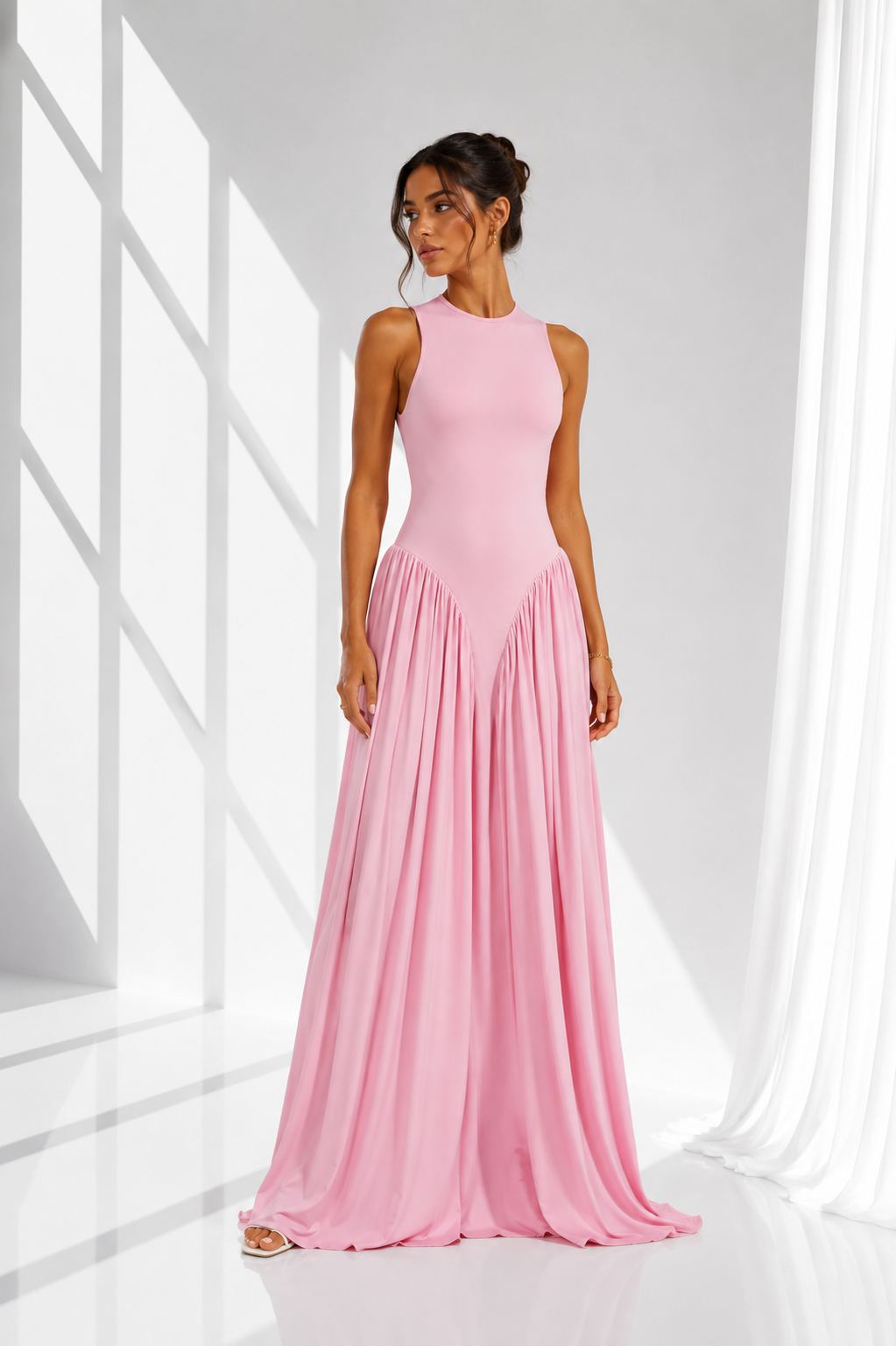 A woman modeling a sleeveless, high-neck, floor-length pink gown with a fitted bodice and flowing pleated skirt, standing in a bright white studio with window-like shadows and sheer curtains.