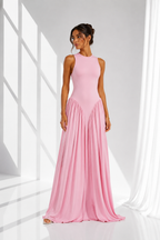 A woman modeling a sleeveless, high-neck, floor-length pink gown with a fitted bodice and flowing pleated skirt, standing in a bright white studio with window-like shadows and sheer curtains.