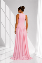 A woman wearing a sleeveless, floor-length pink gown, shown from the back in a bright white studio with window-like shadows.
