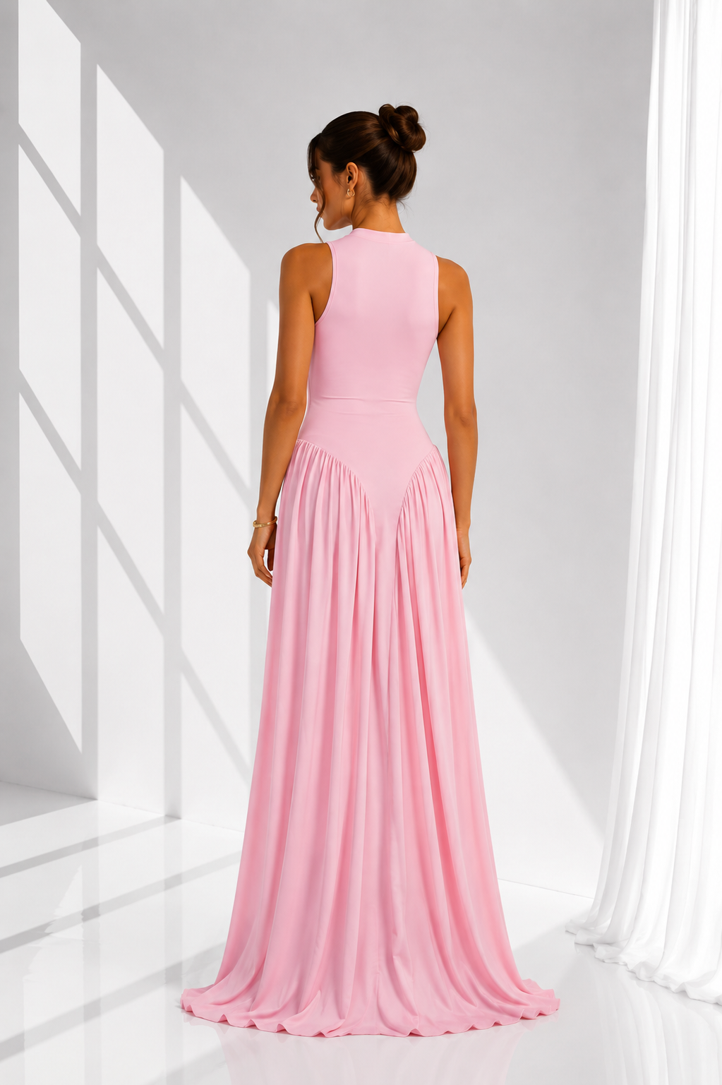 A woman wearing a sleeveless, floor-length pink gown, shown from the back in a bright white studio with window-like shadows.