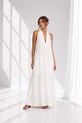 A woman modeling a sleeveless white halter-neck maxi dress with a deep V-neckline, standing in a bright white studio with soft window-like shadows.