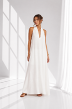 A woman modeling a sleeveless white halter-neck maxi dress with a deep V-neckline, standing in a bright white studio with soft window-like shadows.