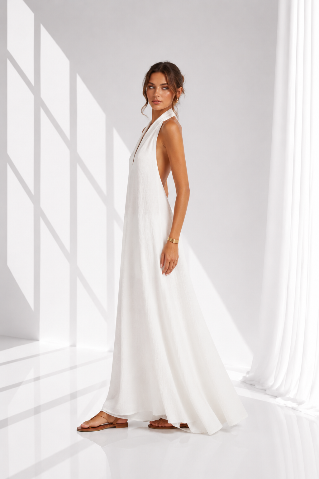 A woman modeling a sleeveless white maxi dress with a deep V-neckline and back, standing in a bright white room with dramatic window shadows.