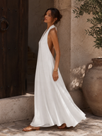 A woman wearing a flowing white sleeveless maxi dress with a deep V-neckline and open back, standing outdoors against a rustic stone wall and wooden door.