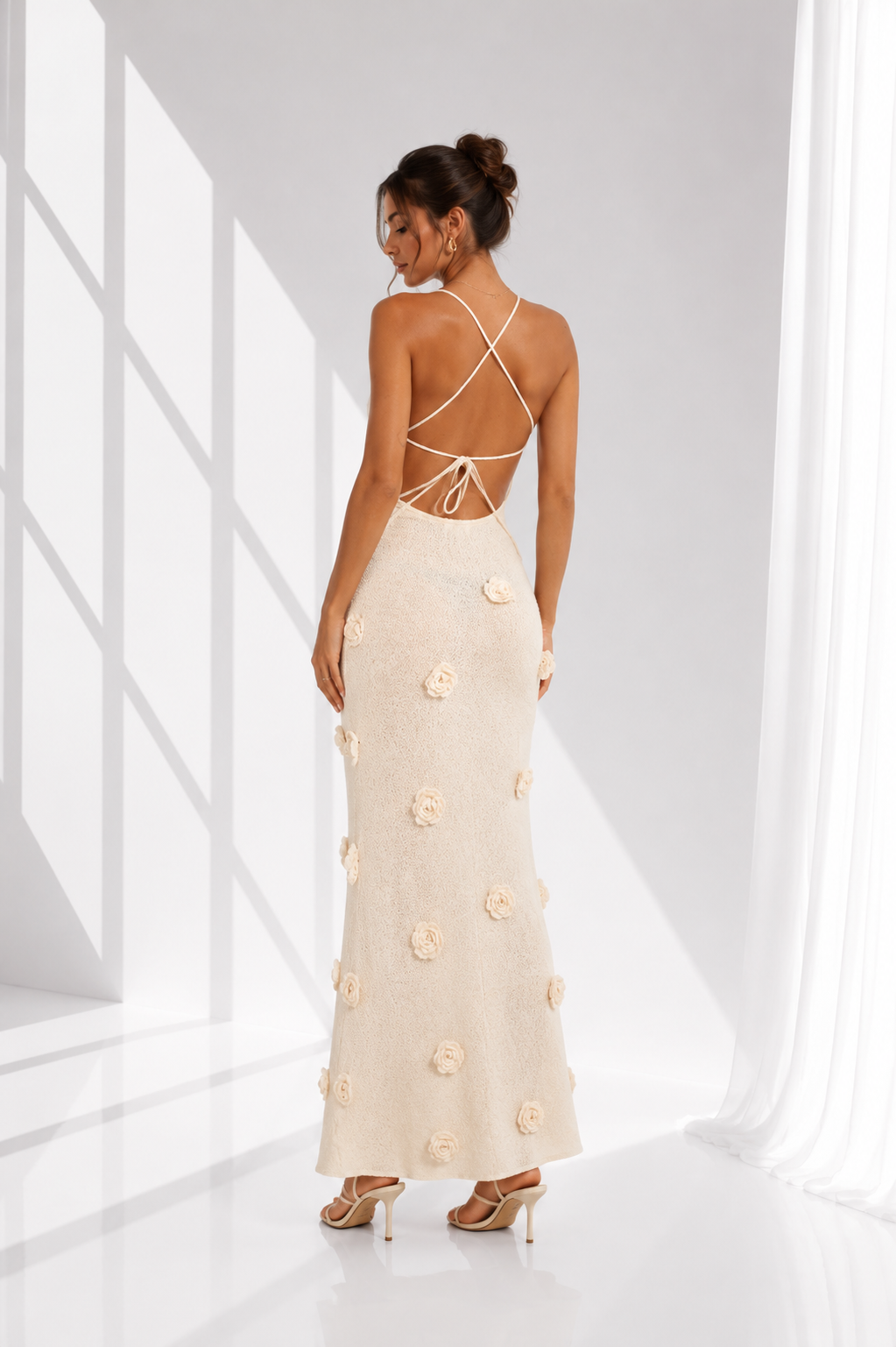 A woman modeling a floor-length cream maxi dress from the back, featuring a strappy open back and 3D floral applique, set against a bright white studio background with window shadow projections.