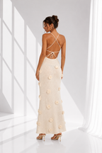 A woman modeling a floor-length cream maxi dress from the back, featuring a strappy open back and 3D floral applique, set against a bright white studio background with window shadow projections.