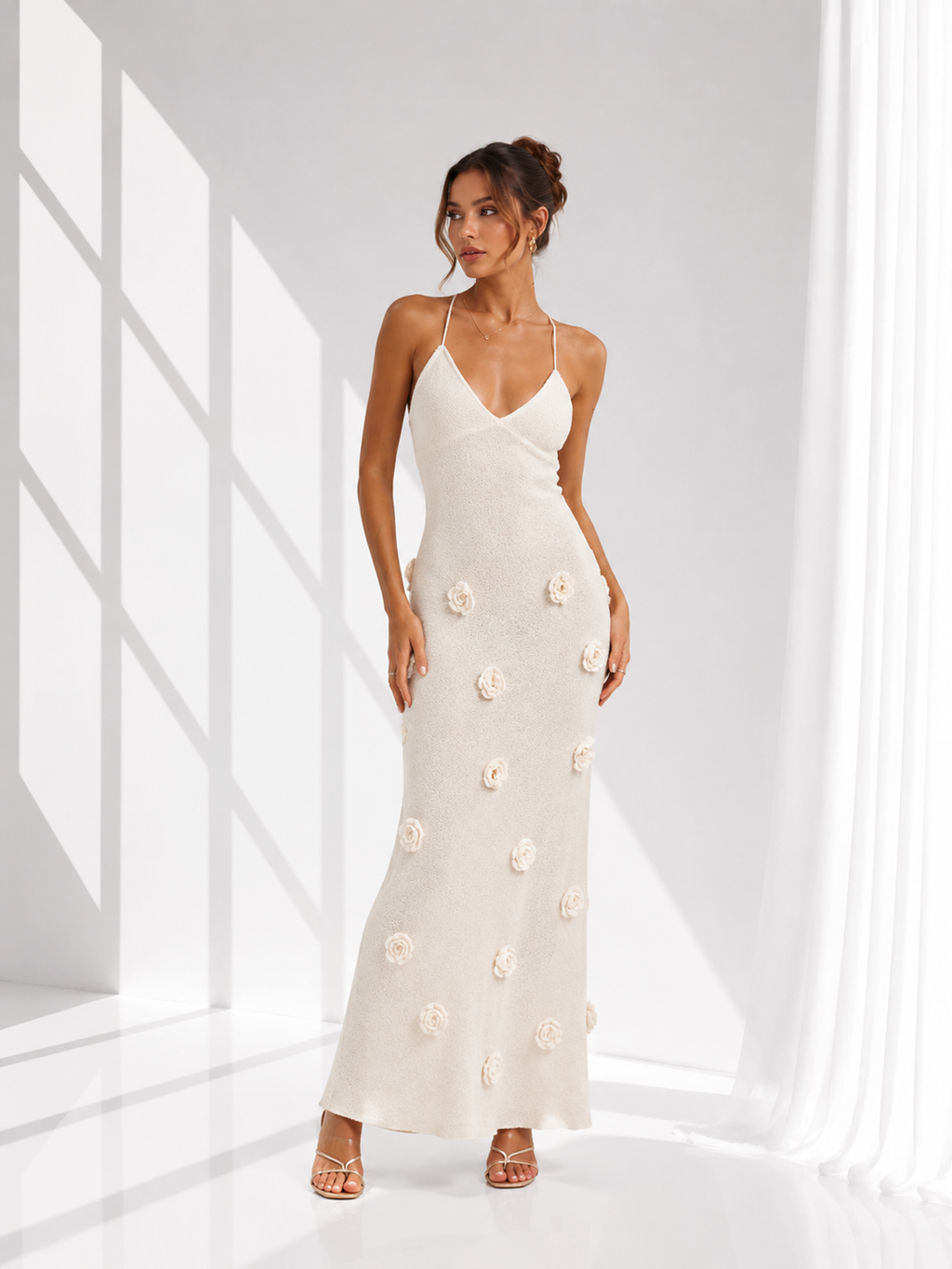 A woman modeling a sleeveless, floor-length cream maxi dress with 3D floral appliques, standing in a bright white room with window shadows.