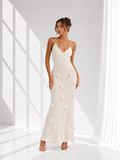 A woman modeling a sleeveless, floor-length cream maxi dress with 3D floral appliques, standing in a bright white room with window shadows.