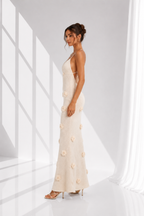 A woman models a floor-length cream or off-white evening gown with 3D floral appliqués, set against a bright white studio background with dramatic window-like shadows.