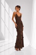 A woman models a dark brown sleeveless maxi dress with thin spaghetti straps and 3D floral appliques, set against a bright white studio background with window pane shadows.