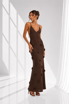 A woman models a dark brown sleeveless maxi dress with thin spaghetti straps and 3D floral appliques, set against a bright white studio background with window pane shadows.