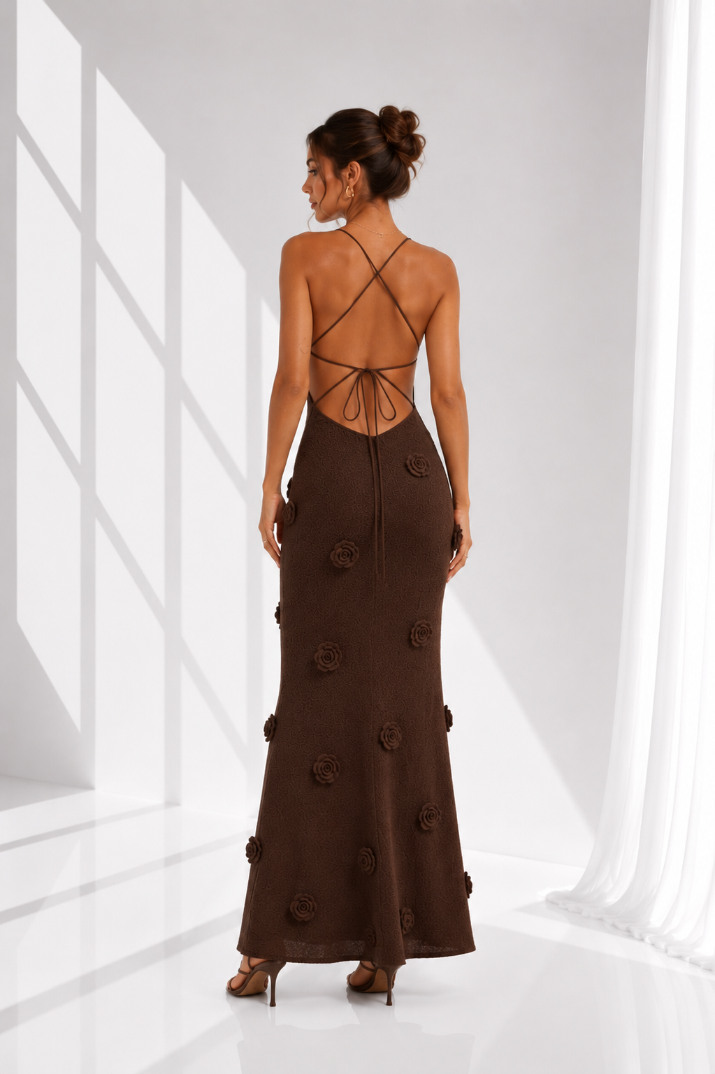 A woman stands with her back to the camera wearing a dark brown floor-length gown with an open back, thin strappy detailing, and raised floral embellishments, photographed in a bright white studio with window-like shadows.