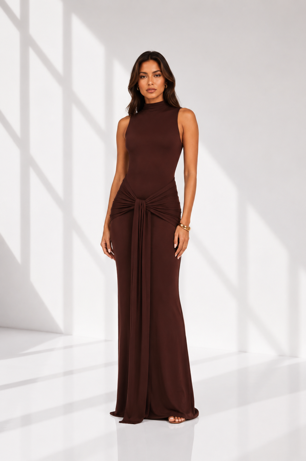 Woman wearing a brown sleeveless dress with a tie waist in a white room.
