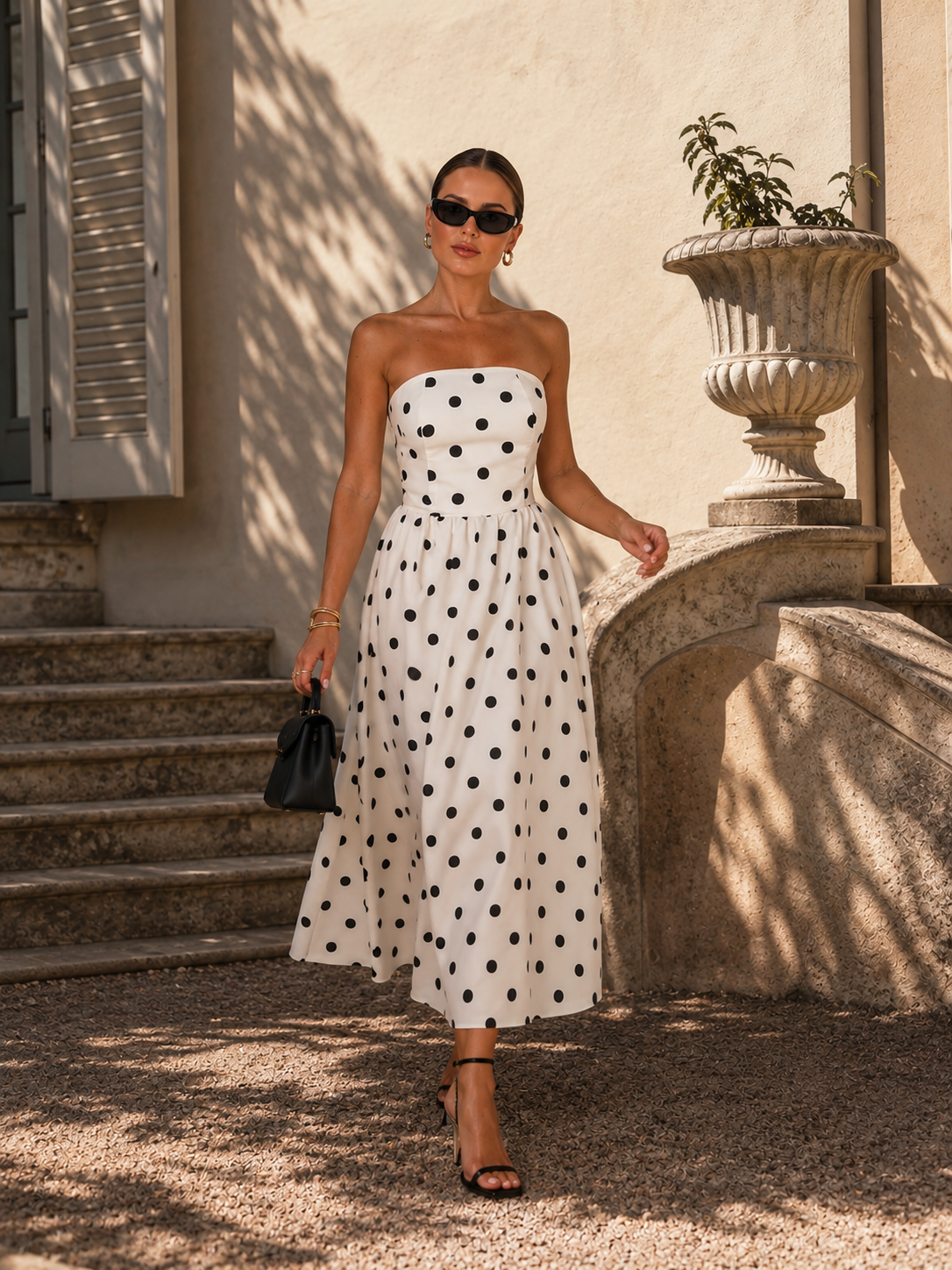 Woman in a strapless polka dot dress standing outdoors near steps and a stone wall.