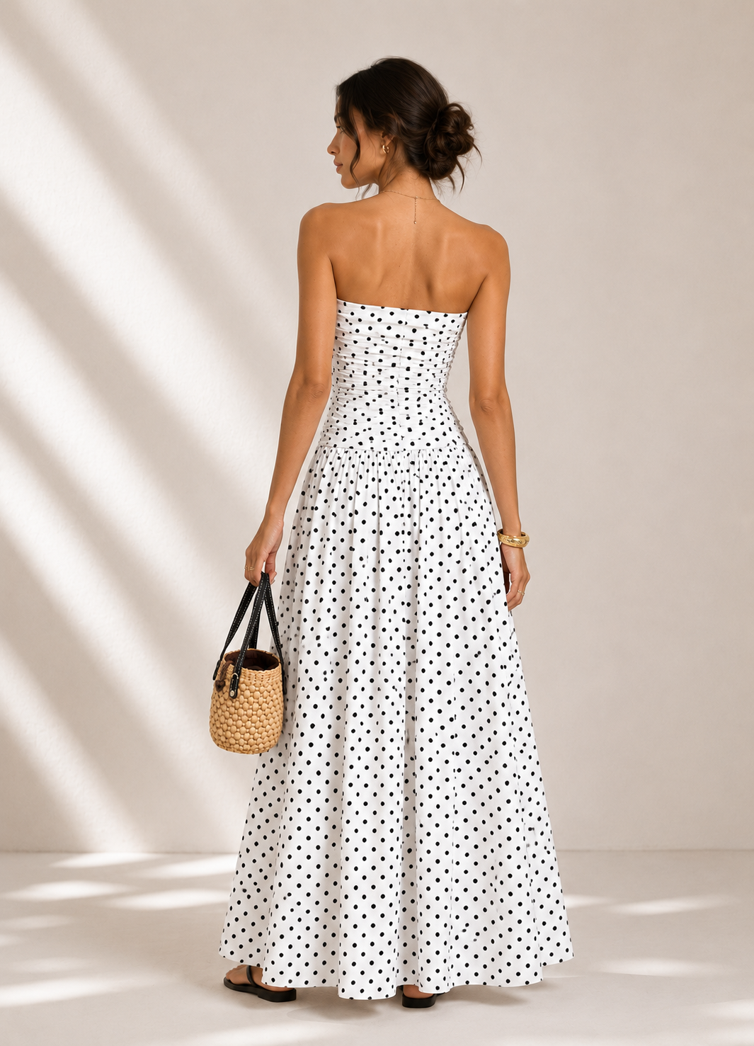 Woman wearing a strapless polka dot dress holding a woven handbag against a neutral background