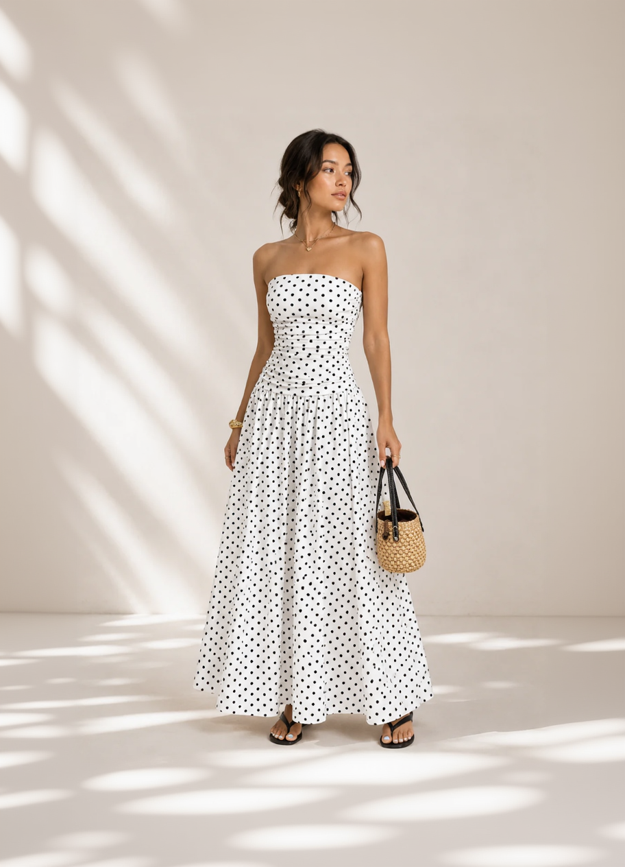Woman wearing a strapless polka dot dress holding a woven handbag against a neutral background
