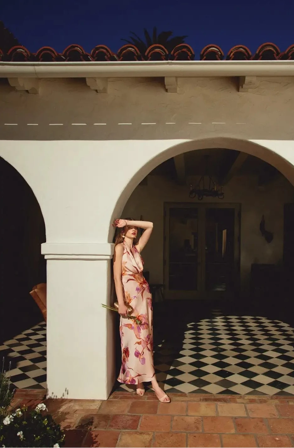 Woman in a floral dress standing in a sunlit archway of a building with a checkered floor.