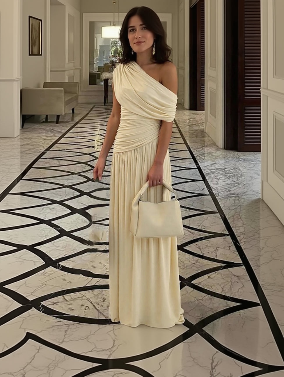 Woman in a flowing beige dress holding a matching handbag in an elegant interior setting.