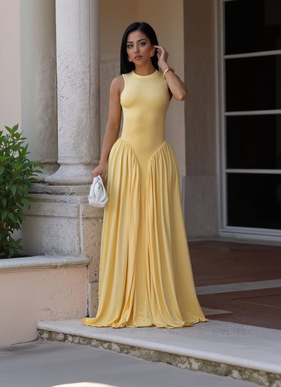 A woman wearing a sleeveless yellow maxi dress with a fitted bodice and flowing skirt, standing outdoors near a stone column and holding a small white handbag.