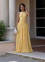 A woman wearing a sleeveless yellow maxi dress with a fitted bodice and flowing skirt, standing outdoors near a stone column and holding a small white handbag.