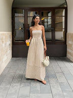 Woman in a long striped dress holding a drink and a bag, standing on a street.