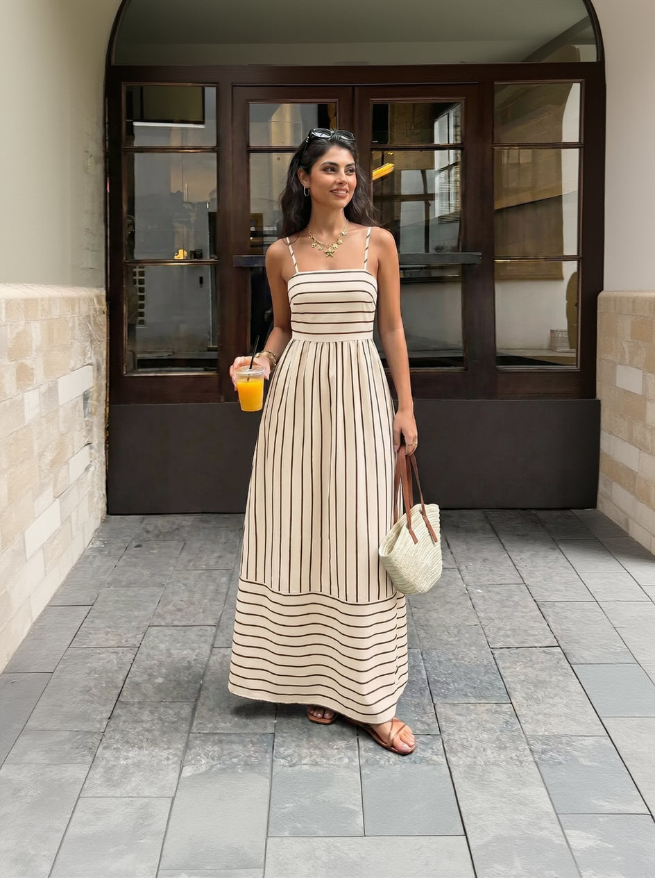 Woman in a long striped dress holding a drink and a bag, standing on a street.