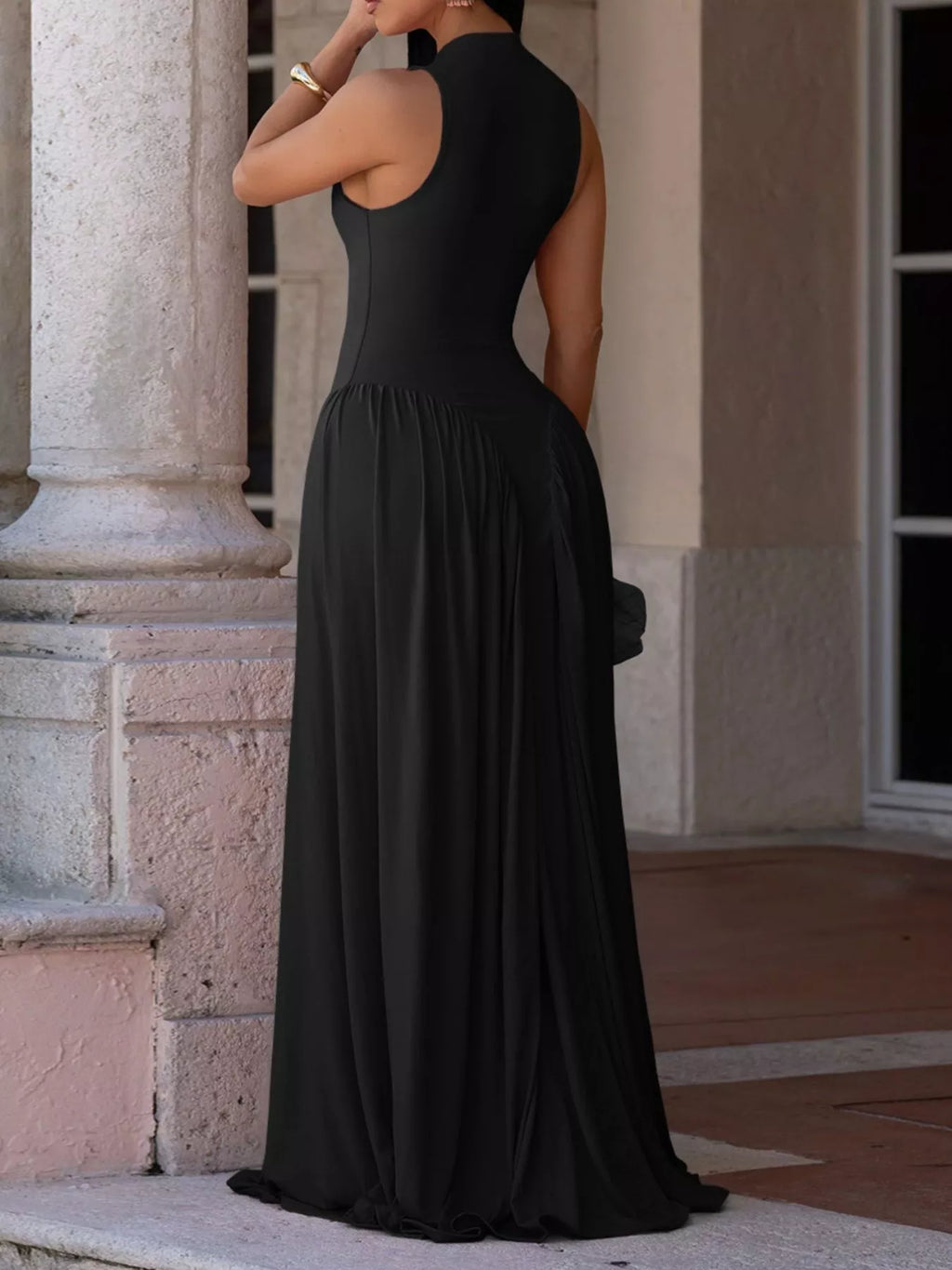 A woman wearing a sleeveless black maxi dress with a high neck and backless open back, standing outdoors in front of a stone building.