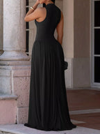 A woman wearing a sleeveless black maxi dress with a high neck and backless open back, standing outdoors in front of a stone building.