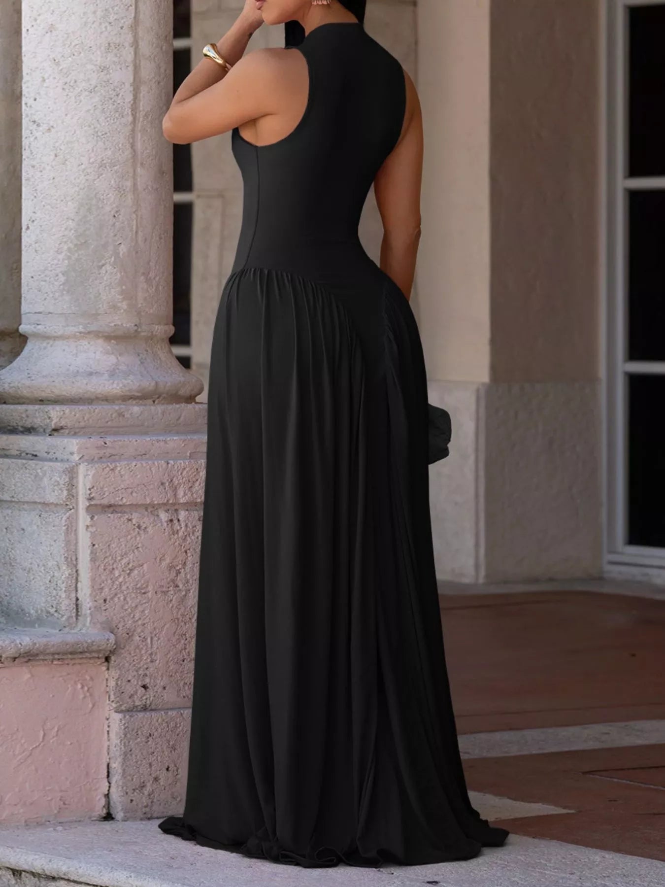 A woman wearing a sleeveless black maxi dress with a high neck and backless open back, standing outdoors in front of a stone building.
